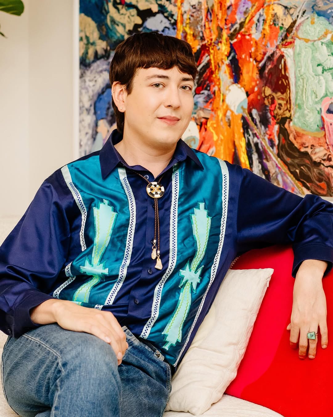Person in blue sitting in chair with a bright coloued painign in the background.